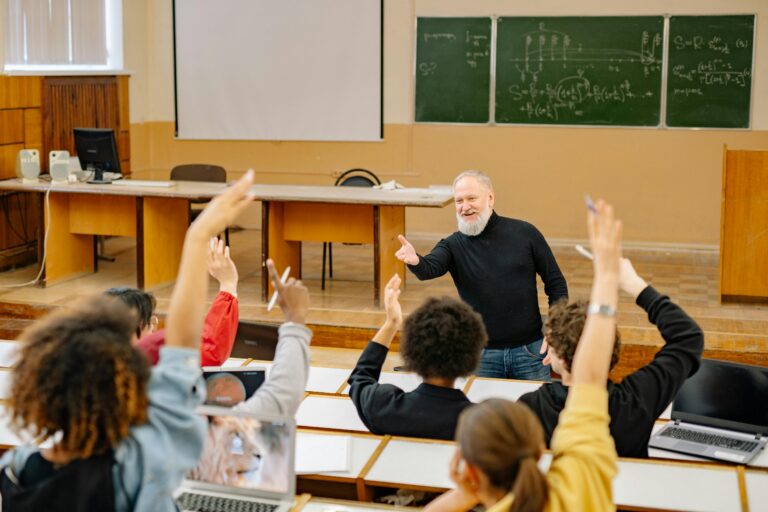 Professor engaging with students in a lively university classroom discussion as students raise hands.